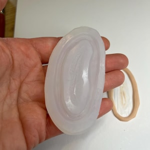 Może przedstawiać: Przezroczysty, owalny kamień z koncentrycznymi pasmami bieli i bladego fioletu. Kamień trzymany jest w dłoni, obok znajduje się inny podobny kamień. Kamienie wydają się być wypolerowane i gładkie.