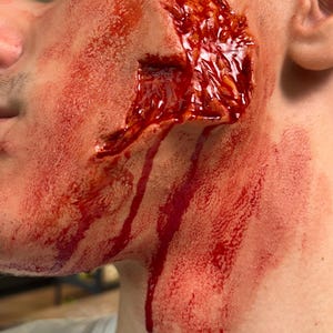 Può includere: Un'applicazione di trucco prostetico su una guancia che presenta una lacerazione profonda e frastagliata con bordi cutanei realistici e testurizzati. La ferita è riempita di una sostanza rosso scuro lucida, con striature di liquido rosso vivo.
