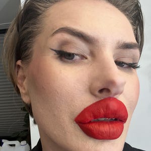 May include: A face featuring bold, matte red lipstick applied to the lips, paired with thin black winged eyeliner on the eyelids. The skin has a natural finish, and the hair is styled back from the forehead, showcasing a clear view of the makeup application.