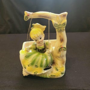 Może przedstawiać: Ceramiczna doniczka w kształcie dziewczynki na huśtawce. Dziewczynka ma na sobie zieloną sukienkę i żółte włosy. Huśtawka jest przymocowana do pnia drzewa z motylem. Doniczka jest ozdobiona żółtymi kwiatami.