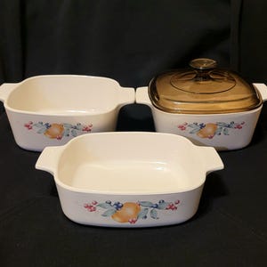 Può includere: Un set di tre teglie da forno in ceramica bianca con un design floreale che presenta frutta e bacche. Le teglie sono quadrate e rettangolari. La teglia più grande ha un coperchio in vetro trasparente.