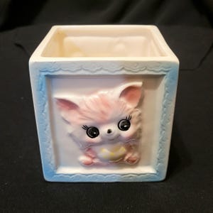 May include: A white ceramic planter with a blue trim and a pink and white cartoon animal face on the front. The animal has large black eyes and a yellow bow.