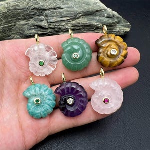 May include: Six gemstone pendants in various colors, including clear, green, and purple, each carved in a spiral seashell design. The pendants have gold-colored bails and small accent stones. The gemstones are held in a hand.