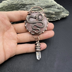 May include: A copper pendant with a rose quartz stone featuring a snake design. A clear quartz crystal dangles from the bottom of the pendant.