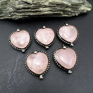 May include: Five rose quartz heart pendants with silver-colored beaded trim. Each pendant has a small loop at the top for a chain. The pendants are displayed on a black surface.
