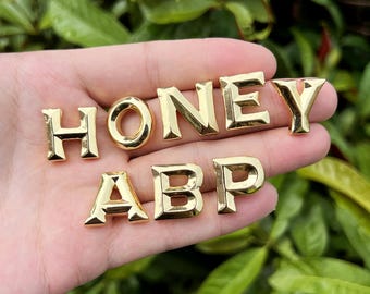 Può includere: Lettere in metallo color oro che compongono le parole "HONEY" e "ABP". Le lettere sono in una mano, con le lettere "HONEY" in alto e "ABP" in basso.