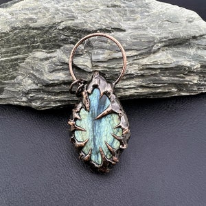 May include: A copper wire wrapped pendant featuring a blue-green labradorite gemstone. The pendant has a rough, organic shape with a wire design that resembles branches or thorns.