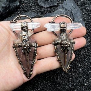 May include: A pair of earrings with arrowhead pendants, clear quartz crystals, and bronze-toned metalwork. The pendants are triangular, with a central design, and are suspended from a circular hoop. The earrings are handcrafted.