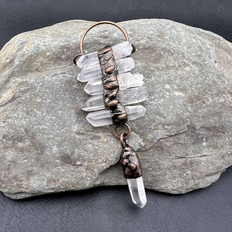 May include: A copper wire wrapped pendant with clear quartz crystals. The pendant is hanging from a rock.
