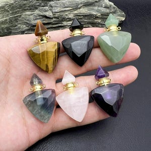 Pode incluir: Seis frascos de perfume de pedras preciosas com detalhes dourados. Os frascos são triangulares com superfícies facetadas e rolhas pontiagudas. As pedras preciosas incluem olho de tigre, ônix preto, aventurina verde, fluorita, quartzo rosa e ametista.