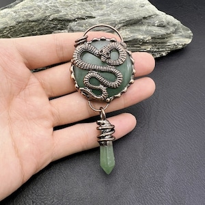 May include: A pendant featuring a green gemstone with a bronze-colored snake design. The pendant has a circular green stone with a coiled snake on top. A green crystal hangs below, wrapped in wire.