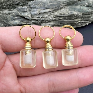 May include: Three small, clear glass perfume bottles with gold-colored accents and a gold ring at the top. Each bottle has a rectangular shape and contains a white interior. The bottles are held in a hand.
