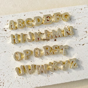 Puede incluir: Abalorios dorados con letras del alfabeto, cada uno incrustado con pequeñas piedras brillantes. Las letras de la A a la Z están dispuestas en filas, mostrando una variedad de fuentes. Los abalorios se exhiben sobre una superficie texturizada de color blanquecino.