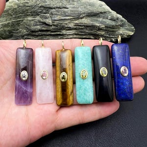 May include: Six rectangular gemstone pendants in shades of purple, pink, brown, teal, black, and blue. Each pendant has a gold-colored bail and a small, oval-shaped gemstone in the center. The pendants are displayed against a dark background.