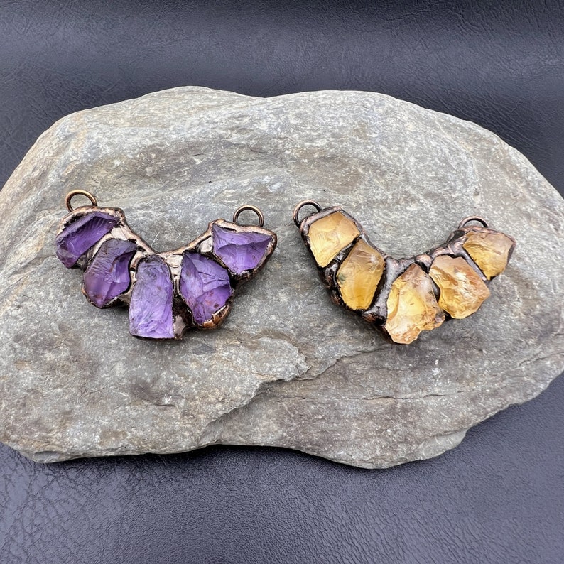 May include: Two copper and gemstone pendants, one with purple amethyst stones and the other with yellow citrine stones. Both pendants are shaped like a crescent moon.