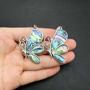 Puede incluir: Dos colgantes de mariposa iridiscentes con detalles plateados. Las mariposas tienen un diseño de concha de abulón azul, verde y rosa.