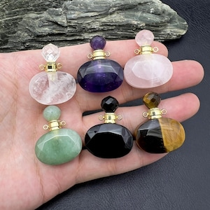 Op de afbeelding: Zes edelsteen parfumflesjes in verschillende kleuren, waaronder transparant, paars, roze, groen, zwart en bruin. Elk flesje heeft een goudkleurige metalen kraag en stopper. De flesjes zijn ovaal van vorm en lijken van gepolijste steen te zijn.