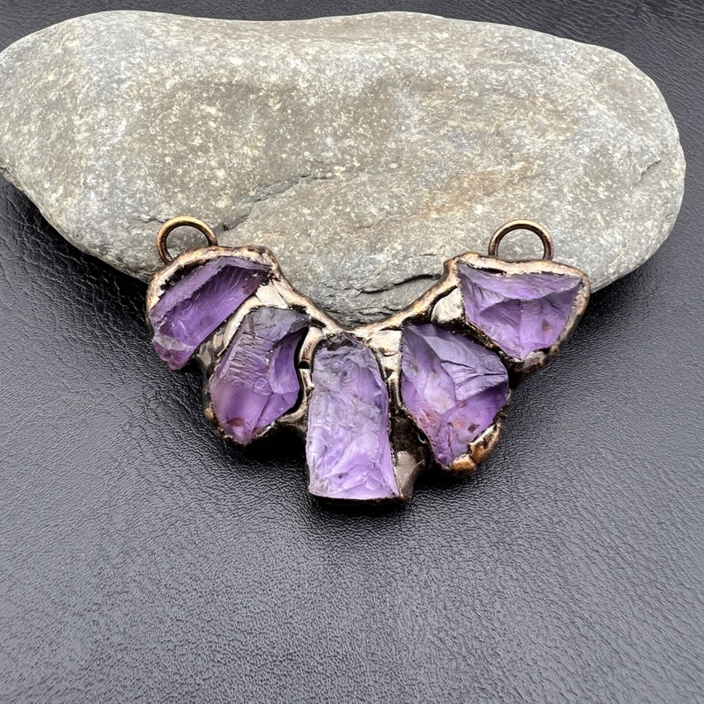 May include: A purple amethyst pendant necklace with a raw, natural crystal design. The pendant is made of five amethyst stones set in a bronze metal frame.