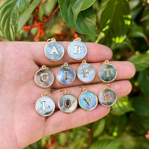 Könnte beinhalten: Eine Sammlung goldfarbener Initialen-Anhänger mit schillerndem Perlmutt-Hintergrund. Jeder Anhänger zeigt einen anderen Buchstaben, darunter A, B, G, I, F, T, L, O, V und E. Die Anhänger sind rund und haben oben eine kleine Schlaufe zum Befestigen an einer Kette.