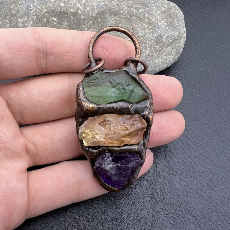 May include: A copper pendant with three different colored gemstones. The top stone is green, the middle stone is yellow, and the bottom stone is purple.