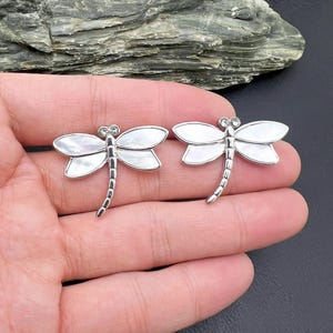 May include: Two silver dragonfly brooches with mother-of-pearl wings and rhinestone accents. The brooches are detailed with a segmented body and delicate antennae. The wings are a pearlescent white, and the brooches are set against a dark background.