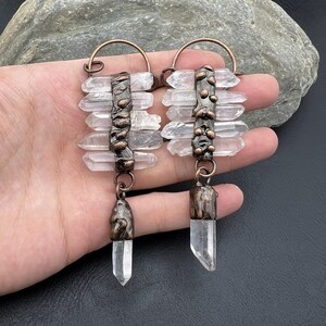 May include: A pair of copper and clear quartz crystal earrings. Each earring features a cluster of quartz crystals attached to a copper wire with a textured copper pendant at the bottom.