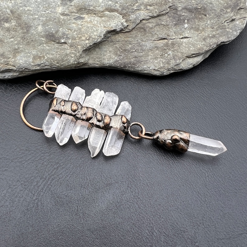 May include: A copper and clear quartz crystal pendant necklace. The pendant features a cluster of quartz points attached to a copper band with a textured surface. The pendant is attached to a copper chain with a clear quartz point at the end.