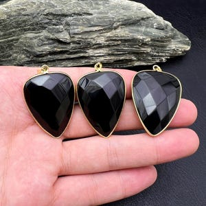 May include: Three black, teardrop-shaped pendants with faceted surfaces and gold-coloured metal trim. The pendants are displayed against a dark background, showcasing their polished appearance and geometric design. The pendants are held in a hand.