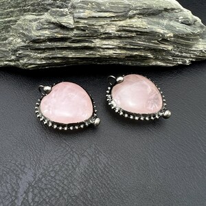 May include: Two heart-shaped rose quartz pendants with a silver-colored beaded border. Each pendant has a small loop at the top for hanging. The pendants are displayed on a black surface.