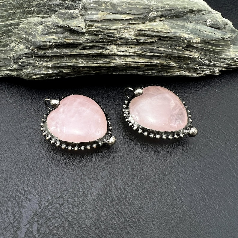 May include: Two heart-shaped rose quartz pendants with a silver-colored beaded border. Each pendant has a small loop at the top for hanging. The pendants are displayed on a black surface.