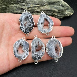 Op de afbeelding: Zes natuurlijke geode hangers met zilverkleurige metalen rand. De geodes zijn voornamelijk grijs en wit met donkergrijze en bruine kristalformaties aan de binnenkant. Elke hanger heeft een lus aan de boven- en onderkant.