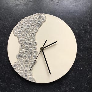 May include: A circular, cream-coloured wall clock with black hands. The left side of the clock face features a decorative pattern of small, white, circular shapes. The clock is mounted on a dark, speckled surface.