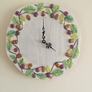 May include: A white ceramic clock with a woodgrain design. The clock face is surrounded by a border of green leaves, brown acorns, and purple berries.