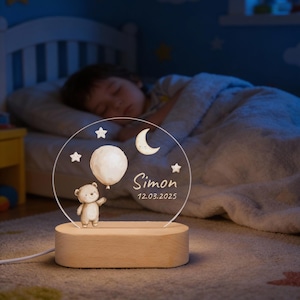 Könnte beinhalten: Personalisiertes Nachtlicht mit einem Bären, der einen Ballon, Sterne und einen Halbmond hält. Der Name "Simon" und das Datum 12.03.2025 sind abgebildet. Das Licht steht auf einem rechteckigen Holzsockel, mit einem schlafenden Kind im Hintergrund.