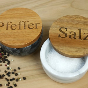 Könnte beinhalten: Zwei Marmorsalz- und Pfefferstreuer mit Holzdeckeln. Die Deckel sind mit den Worten "Pfeffer" und "Salz" graviert.