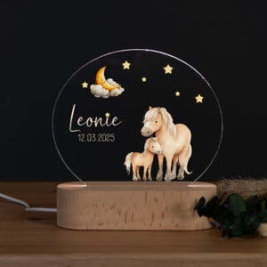 Könnte beinhalten: Personalisiertes Nachtlicht mit kreisförmigem Acryldesign. Es zeigt eine Pferde-Familien-Illustration, einen Mond, Sterne und den Namen "Leonie" mit dem Datum 12.03.2025. Das Licht steht auf einem Holzsockel.
