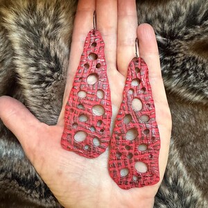 May include: A pair of long, red leather earrings with a geometric, openwork design. The earrings are teardrop shaped and have a textured surface.