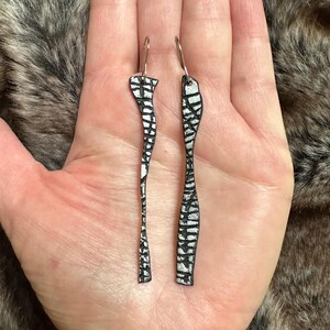 May include: A pair of long, black and silver earrings with a geometric pattern. The earrings are made of a textured material and have a dangling design.