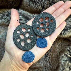 May include: Black and blue earrings. The earrings have a textured, circular design with multiple holes, and a smaller, textured blue pendant. The earrings are held in a hand against a furry background.