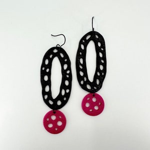 May include: A pair of black dangle earrings with a pink circle pendant. The earrings are made of a lightweight material with a cutout design.