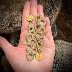 May include: Gold-toned earrings with a textured, wavy design and circular cutouts. Each earring features a dangling circular charm.