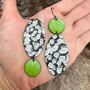 May include: A pair of dangle earrings featuring a black and white oval design with a green circular pendant. The earrings are made of metal and have a textured finish.