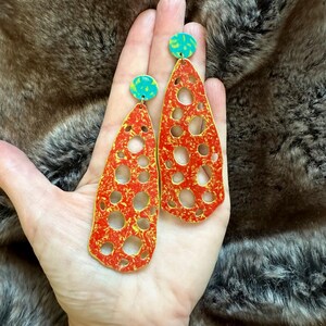 May include: A pair of long, red and gold earrings with a cutout design. The earrings have a turquoise blue accent at the top.