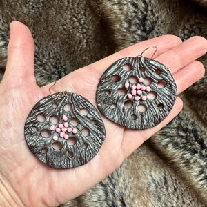 May include: A pair of dark brown, circular earrings with a textured surface and numerous holes. Small pink beads are clustered in the centre of each earring. The earrings are held in a hand against a furry background.