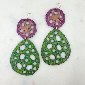 May include: A pair of dangle earrings with a green and purple iridescent finish. The earrings feature a round top piece with a circular cutout pattern and a teardrop-shaped bottom piece with a similar cutout pattern.