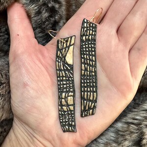 May include: A pair of long, black and gold earrings with a geometric pattern. The earrings are made of clay and have a textured surface.