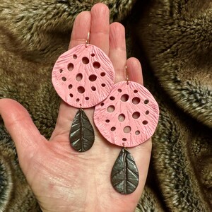 May include: A pair of pink and brown dangle earrings. The earrings are made of polymer clay and feature a round, openwork design with a teardrop-shaped pendant.