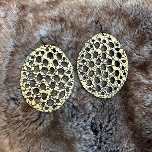 May include: A pair of large, gold-toned earrings with a circular, openwork design. The earrings have a textured, almost honeycomb-like pattern.