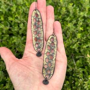 May include: A pair of long, gold and black earrings with a cutout design. The earrings are oval shaped and have a dangling disc at the bottom.
