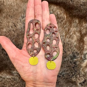 May include: A pair of dangle earrings with a unique, abstract design. The earrings are made of metal and have a textured, multi-colored finish. The earrings are accented with small, round, yellow charms.
